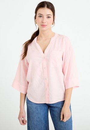 Młoda kobieta z długimi brązowymi włosami, nosząca złote kolczyki-koła, luźną bluzkę na guziki w jasno różowym kolorze oraz niebieskie dżinsy, na białym tle.