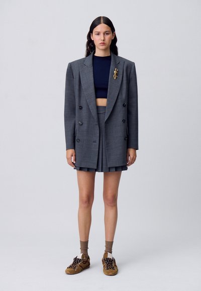 Blazer gris de doble botonadura con solapas anchas, combinado con una mini falda plisada a juego. Debajo, un top corto azul marino y zapatillas marrones.