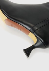 Fekete bőr magassarkú cipő hegyes orral, sima felülettel és vékony, döntött sarokkal. Alatta világosbarna bőr talpbetét található.