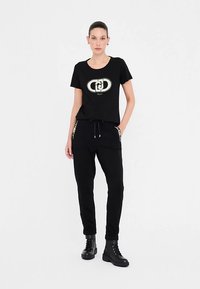 Sort T-shirt med et metallisk logo, kombineret med sorte joggers med gyldne detaljer på lommerne og snøre i taljen. Stylet med sorte støvler.