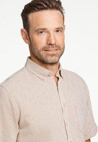 Jasnobeżowa koszula na krótkie rękawy zapinana na guziki, wykonana z fakturowanej tkaniny, z pojedynczą kieszenią na piersi oraz kołnierzykiem ozdobionym guzikami.