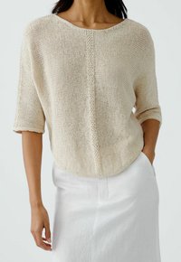 Top tricot beige con vestibilità ampia e scollatura a barca; presenta maniche corte e una cucitura centrale texturizzata; indossato con una gonna bianca.