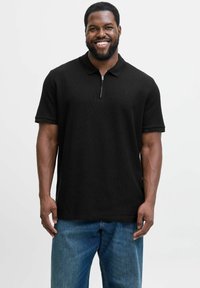 Sort polo skorte med halvlukket krave, korte ærmer og et struktureret stof, parret med blå løstsiddende jeans.