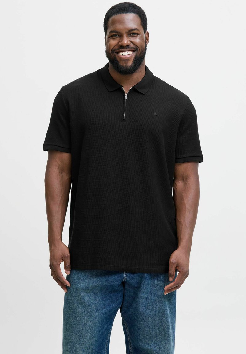 Schwarzes Poloshirt mit einem halben Reißverschlusskragen, kurzen Ärmeln und einem strukturierten Stoff, kombiniert mit blauen, locker sitzenden Jeans.