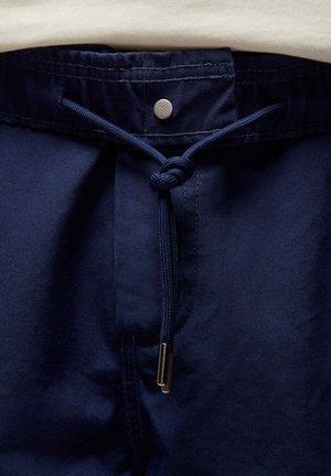 Marineblauwe broek met een trekkoord in de taille, zilverkleurige hardware en een gestructureerde stof. Heeft een knoopdetail op het trekkoord.