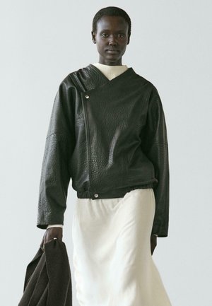 Osoba s tmavou pletí v texturované černé kožené bundě přes bílé šaty, držící v jedné ruce tmavý kabát, stojící před jednobarevným pozadím.