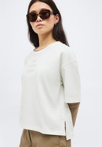 Γυναίκα με μακριά σκουρόχρωμα μαλλιά που φορά γυαλιά ηλίου με καφέ σκελετό τύπου χελώνας, μπεζ φαρδιά μπλούζα με το κείμενο «ΤΟ ΜΕΛΛΟΝ ΕΙΝΑΙ ΔΙΚΟ ΜΑΣ» και καφέ παντελόνι.