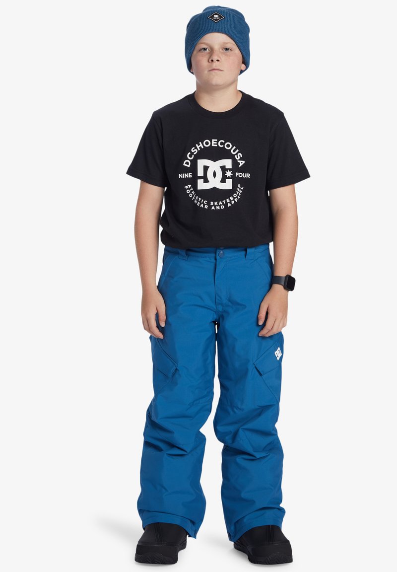 Blå snöbyxor med flera fickor, bärs med en svart t-shirt som har en vit logotyp, och en blå stickad mössa. Svarta skor fulländar outfiten.