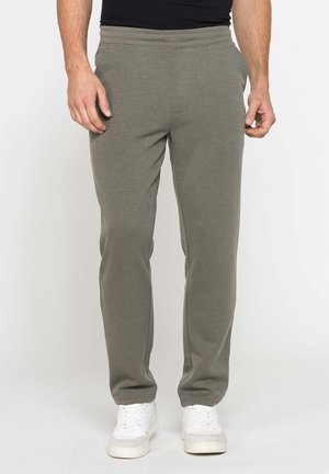 Uomo con pantaloni della tuta verde oliva e sneakers bianche, in piedi davanti a uno sfondo semplice con le mani lungo i fianchi.