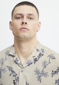 Camisa de manga corta beige con patrones florales azules, con diseño de cuello y detalles de botones en la parte delantera.
