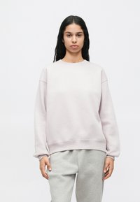 Γυναίκα ντυμένη με ροζ φούτερ και γκρίζες φόρμες, σε απλή λευκή/background, στραμμένη προς τα εμπρός με ουδέτερη έκφραση.