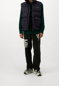 Gilet imbottito blu sopra una camicia verde, pantaloni neri con toppe grafiche bianche e sneakers in bianco e nero. Testura liscia e design casual.