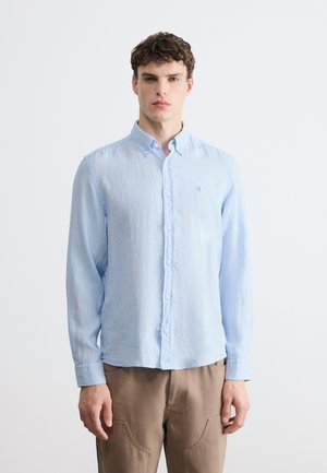 Νεαρός άνδρας με κοντά σγουρά μαλλιά, που φοράει ανοιχτό μπλε πουκάμισο με κουμπιά και μακρυμάνικο και μπεζ παντελόνι, στέκεται μπροστά σε απλό λευκό φόντο.