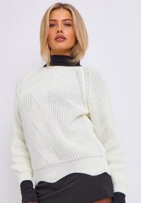 Maglione leggero color crema, caratterizzato da un design a treccia testurizzata, maniche ampie e orlo a coste. Indossato sopra un dolcevita nero.