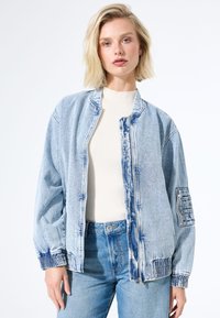 Femme blonde portant une veste bomber en denim bleu clair avec une poche à fermeture éclair sur la manche, sur un haut à côtes blanc et un jean bleu taille haute.