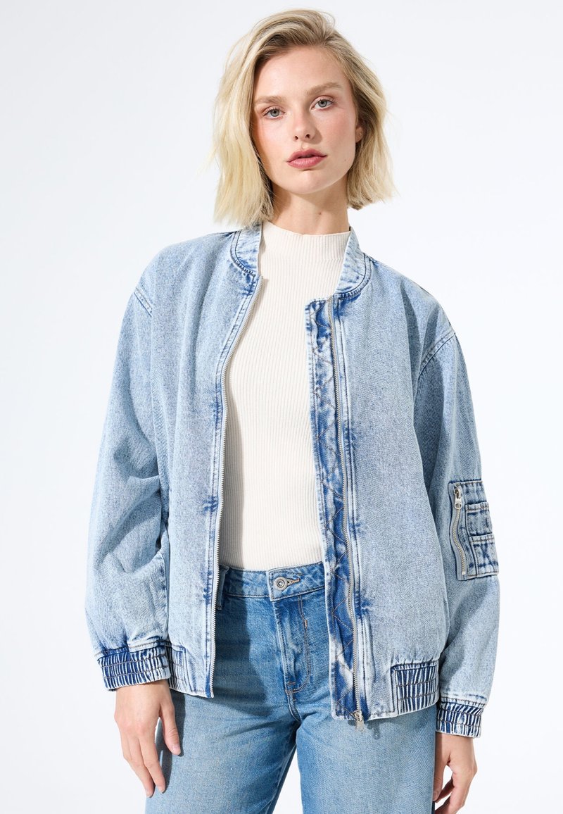 Femme blonde portant une veste bomber en denim bleu clair avec une poche à fermeture éclair sur la manche, sur un haut à côtes blanc et un jean bleu taille haute.