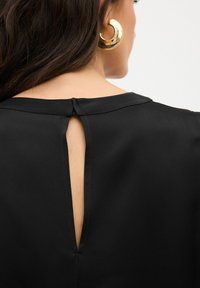 Blusa nera con dettaglio a chiave sulla schiena, realizzata in tessuto liscio. Presenta un colletto discreto e cuciture minime sul collo.