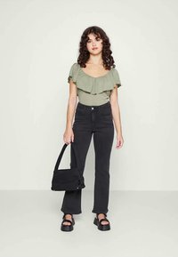 Mulher com cabelo encaracolado, vestindo uma blusa verde-oliva com babados, calças pretas de cintura alta, sandálias de plataforma, segurando uma bolsa preta ao seu lado.