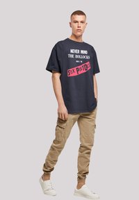 Navy cotton t-shirt with white and pink text, featuring the phrase "NEVER MIND THE BOLLOCKS HERE'S THE SEX PISTOLS." Casual fit, paired with beige cargo pants.