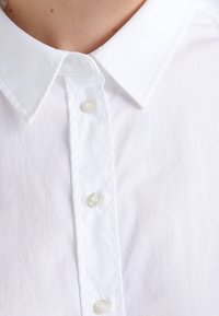 Camicia bianca con colletto classico, texture liscia e cinque bottoni perlacei lungo la pattina anteriore.