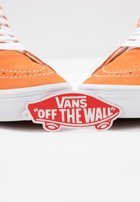 Chaussures Vans en daim orange avec des lacets blancs, une semelle blanche et une étiquette logo rouge "Off the Wall" en forme de planche de skateboard.
