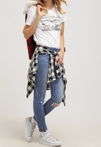 Camiseta gráfica blanca, camisa de cuadros en blanco y negro atada a la cintura, jeans azul desgastados y zapatillas grises claras con tres rayas.