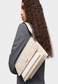 Bolso color crema con un patrón floral texturizado, con un diseño en capas y un logo dorado. Lleva un strap desmontable para usar en el hombro.