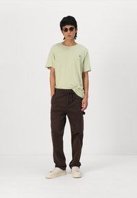 Light green t-shirt, loose fit with short sleeves, paired with brown cargo pants featuring pockets. Model also wears round sunglasses and light sneakers.