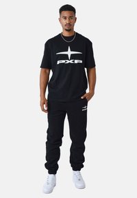 Camiseta negra con gran logotipo blanco "PXP" y gráfico de estrella. Combinada con pantalones de chándal negros con un pequeño detalle del logotipo. Zapatillas blancas.