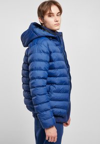 Blå puffjakke med hætte, med pyntet design og blank overflade, lynlås lukning og sidelommer. Modellen har blå bukser på.