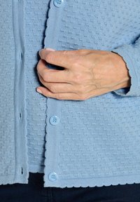 Hellblauer Strickcardigan mit strukturierter Oberfläche, gewellten Kanten und großen blauen Knöpfen, ausgestattet mit einer durchgehenden Knopfleiste und eng anliegenden Bündchen.