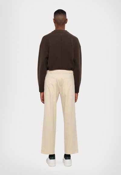 Maglione marrone a maglia con vestibilità rilassata e scollo rotondo, abbinato a pantaloni larghi crema. Sneakers bianche visibili nella parte inferiore.
