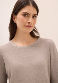 Hellgrauer, strukturierter Pullover mit Rundhalsausschnitt und langen Ärmeln, getragen von einer Person mit dunklem Haar und Sommersprossen, vor einem neutralen Hintergrund.