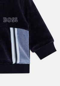 Sweatshirt en velours bleu marine avec des accents gris, logo "BOSS" brodé et détails rayés en bleu clair et bleu foncé, fini avec des poignets côtelés.