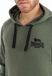 Sudadera color verde oliva hecha de una mezcla de algodón. Presenta cordones negros, un logo de león bordado y el texto "Lonsdale London" en el pecho.
