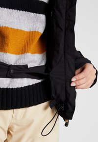 Veste imperméable noire avec fermeture à pression et cordon de serrage, portée par-dessus un pull en tricot rayé multicolore jaune, gris et noir.
