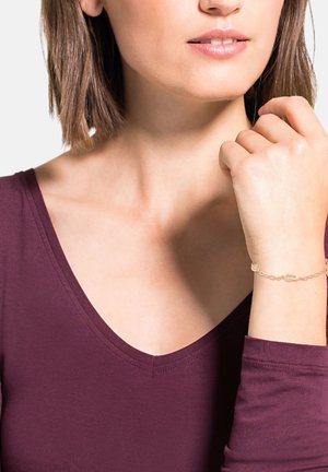 Pulsera de oro con eslabones ovalados, colocada en una muñeca, que muestra un diseño de cadena fina. Top de cuello en V de tela burdeos oscura.
