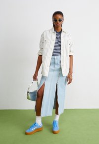 Jupe en denim avec fentes sur les côtés, associée à une chemise rayée et une chemise blanche à manches amples. Baskets bleu clair avec semelles en caoutchouc et sac à main métallique.