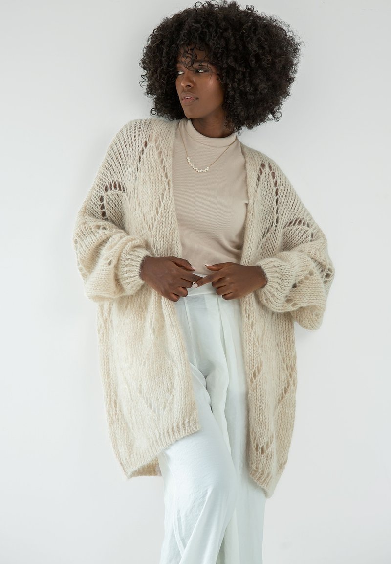 Gruba, kremowa kardigan z dzianiny, z otwartym przodem, złożonymi wzorami koronkowymi i opadającymi ramionami. Noszony na beżową bluzkę i jasne spodnie.
