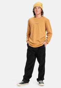 Vaatteet: pitkähihainen sinapinkeltainen paita logolla, yhdistettynä mustiin corduroy-housuihin ja mustiin lenkkareihin. Asusteena ribattu keltainen beanie.