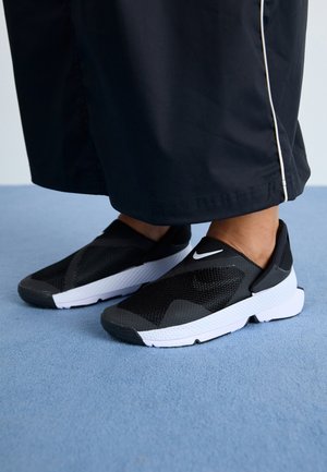 Pieds portant des baskets Nike noires sans lacets avec des semelles blanches, debout sur un tapis bleu, avec un pantalon noir présentant des bandes blanches sur les côtés.
