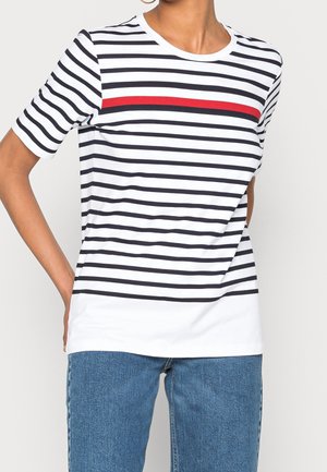Femme portant une chemise blanche à rayures noires avec une seule rayure rouge au niveau de la poitrine et un jean en denim bleu, debout avec une main sur la hanche.