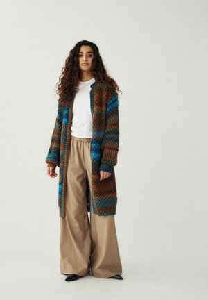 Femme aux longs cheveux bouclés portant un cardigan en tricot multicolore, une chemise blanche, un pantalon large beige et des chaussures noires, debout devant un fond uni.