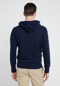 Muž v námořnické modré mikině a béžových kalhotách stojí čelem k volnému světlému pozadí.