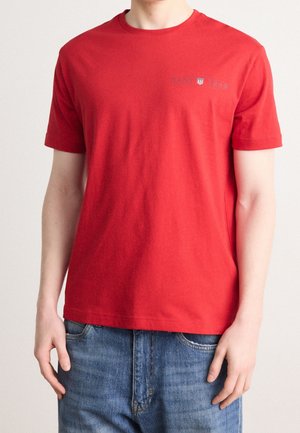 Mand iført en ensfarvet rød kortærmet T-shirt med lille brystlogo kombineret med blå jeans, vist fra hals til hofter.