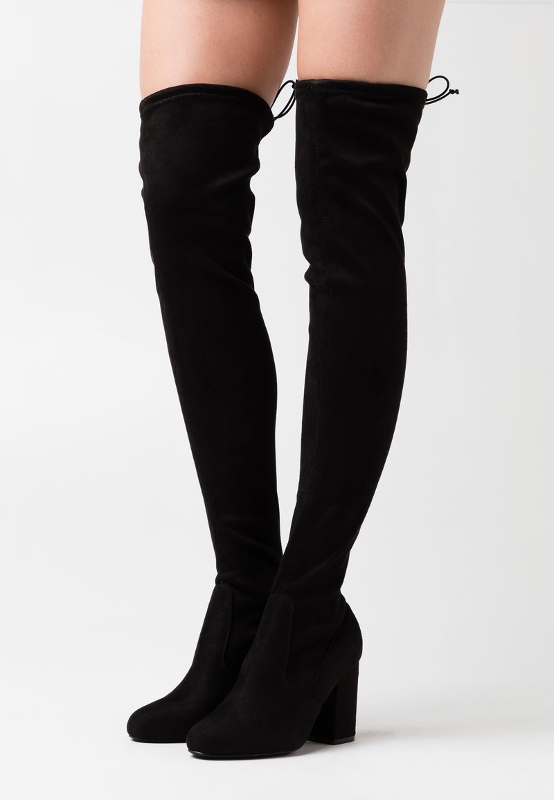 Botas de ante negras hasta el muslo con tacones de bloque, llevadas en las piernas contra un fondo blanco liso.