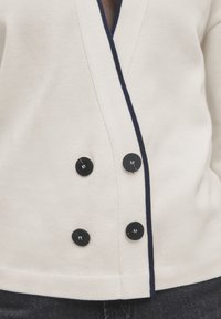 Cream cardigan with a deep V-neck, navy trim, and six black buttons, featuring a textured knit material.