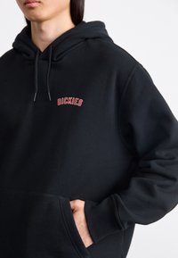 Felpa nera con cappuccio realizzata in tessuto morbido, dotata di polsini e orlo a coste, tasca a marsupio e logo "DICKIES" ricamato in rosso.
