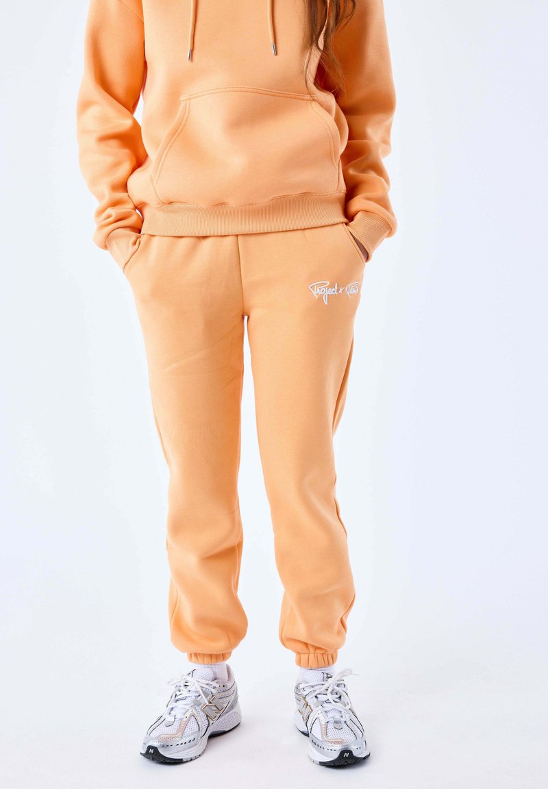 Peachfarvet hættebluse med en frontlomme og matchende joggers; begge har ribbede manchetter og et broderet logo i hvid. Sneakers med mesh-detaljer.