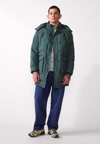 Giacca piumino verde con cappuccio, chiusura a zip e bottoni a pressione, due tasche frontali e texture trapuntata, indossata sopra una maglietta grigia e jeans blu.
